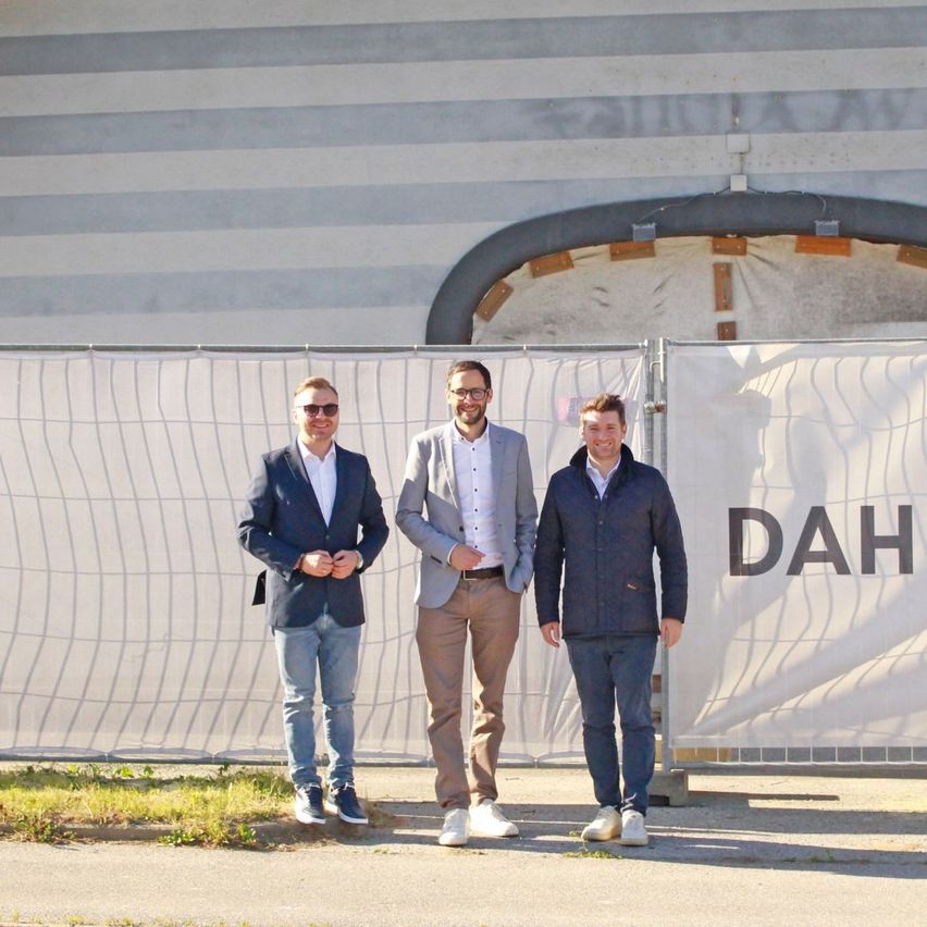 Three men stand outside, smiling, with one wearing glasses. They're in front of a building with 'DAH' on a white wall and a fence.