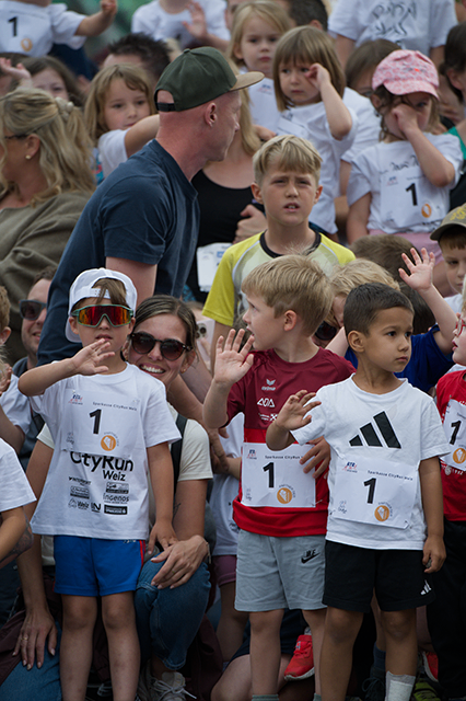 Eine Gruppe von Kindern und Erwachsenen steht in einer Menschenmenge, einige tragen weiße T-Shirts und andere rote. Die Kinder tragen Nummernschilder mit den Nummern 1 und 7.