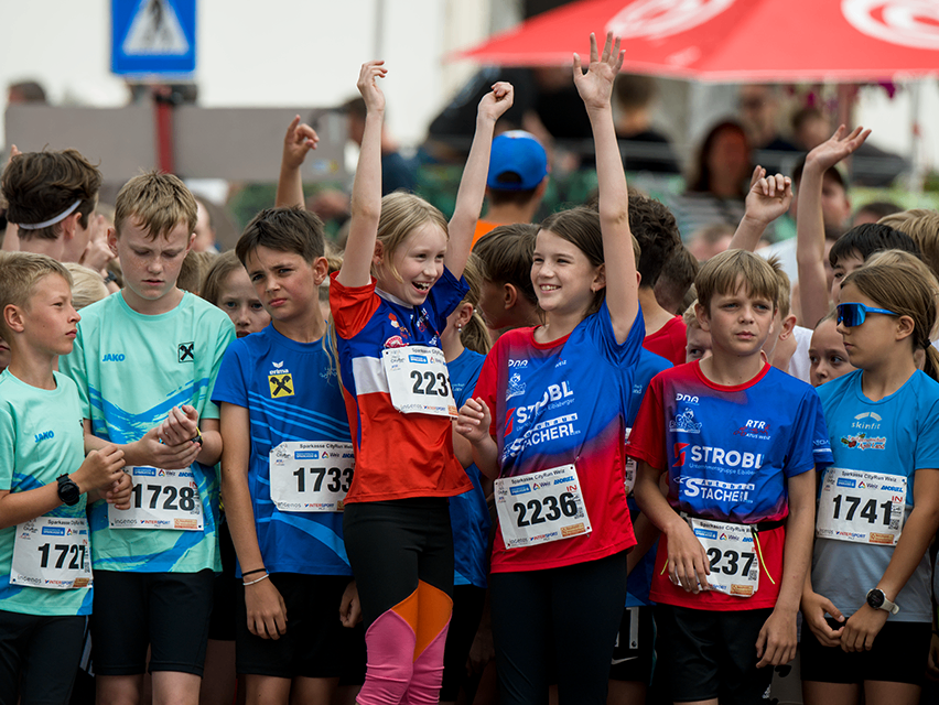 Eine Gruppe von Kindern, alle mit Laufnummern, feuert eine Läuferin mit der Nummer 223 an. Die Menge steht in einer Reihe, viele Hände sind erhoben.