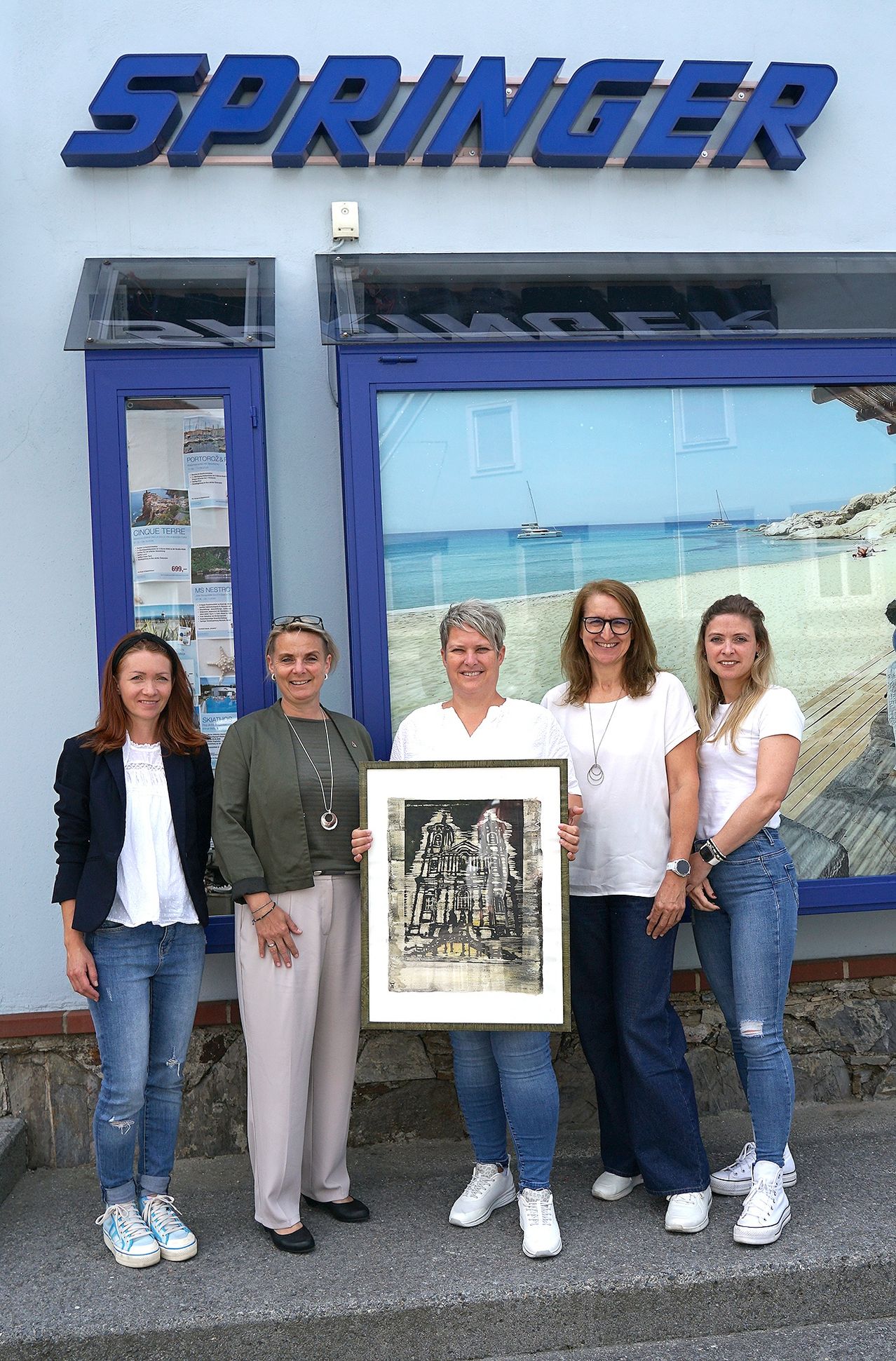 Vier Frauen stehen vor einem Gebäude mit einem gerahmten Kunstwerk in der Hand und lächeln für ein Bild vor einer szenischen Strandanzeige.