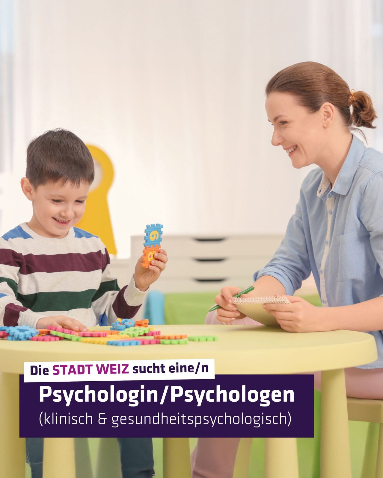 A woman and a boy are sitting at a table, the boy is playing with colorful building blocks while the woman is smiling and holding a notebook and pen. The woman is wearing a blue shirt and the boy is wearing a striped shirt. The background shows a white wall and a yellow object.