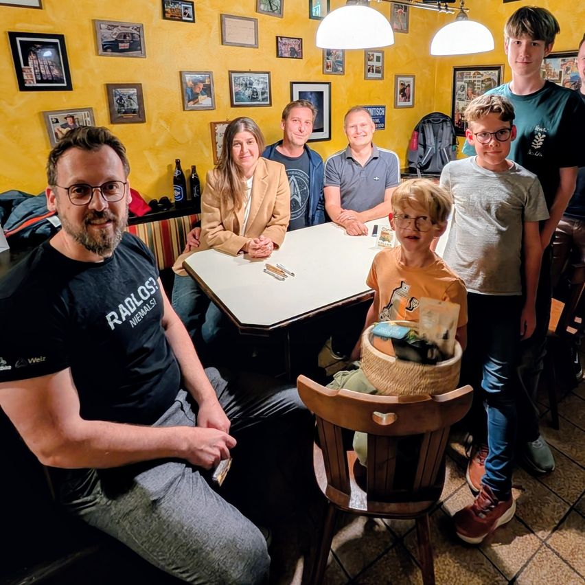 Eine Familie und Freunde versammeln sich um einen Tisch in einem Restaurant, lächeln und posieren für ein Foto. Der Tisch ist mit verschiedenen Gegenständen besetzt, und die Wände sind mit Bilderrahmen dekoriert.