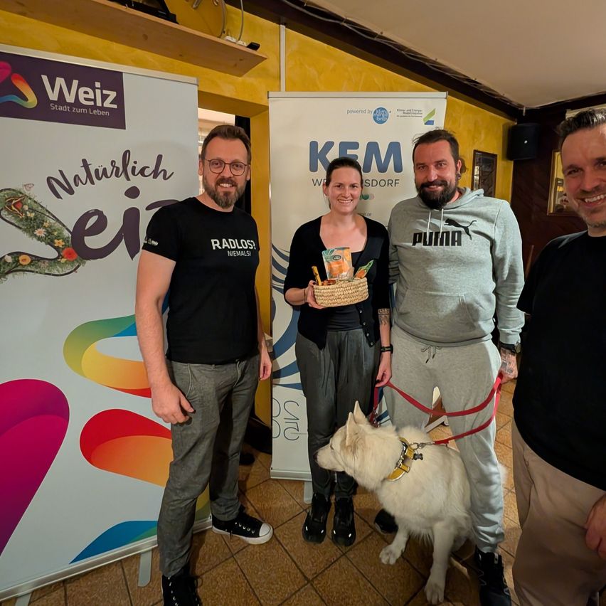 Vier Personen und ein Hund stehen vor einem farbenfrohen Hintergrund mit einem Banner, auf dem 'Naturlich ein' steht. Der Mann links trägt ein schwarzes T-Shirt mit weißem Text, und die Frau in der Mitte hält einen Korb. Der Mann rechts trägt eine graue Kapuzenjacke mit dem Wort 'Puma'.