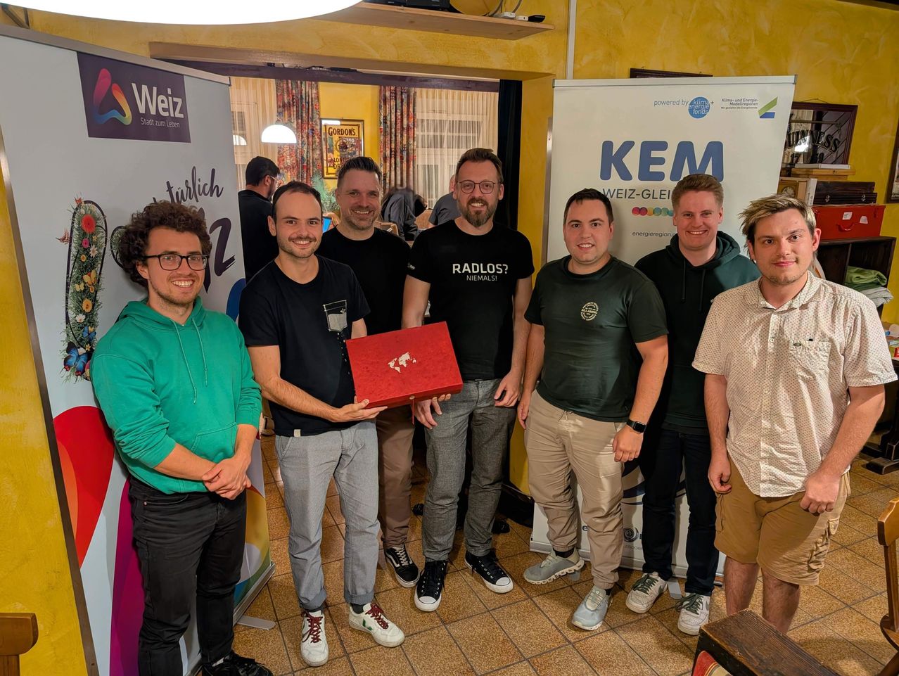 Eine Gruppe von Männern steht lächelnd zusammen, einer hält eine rote Box. Sie stehen vor einer gelben Wand mit Bannern.