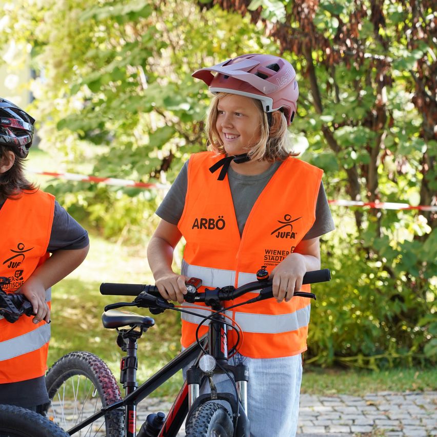 Zwei Kinder stehen auf einem gepflasterten Weg, tragen Sicherheitswesten und Helme. Ein Kind hält ein Fahrrad, und das andere steht neben seinem Fahrrad.