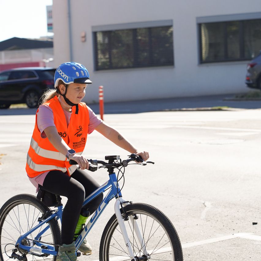 Ein junges Mädchen fährt auf der Straße ein blaues Fahrrad. Sie trägt eine Sicherheitsweste und einen Helm.