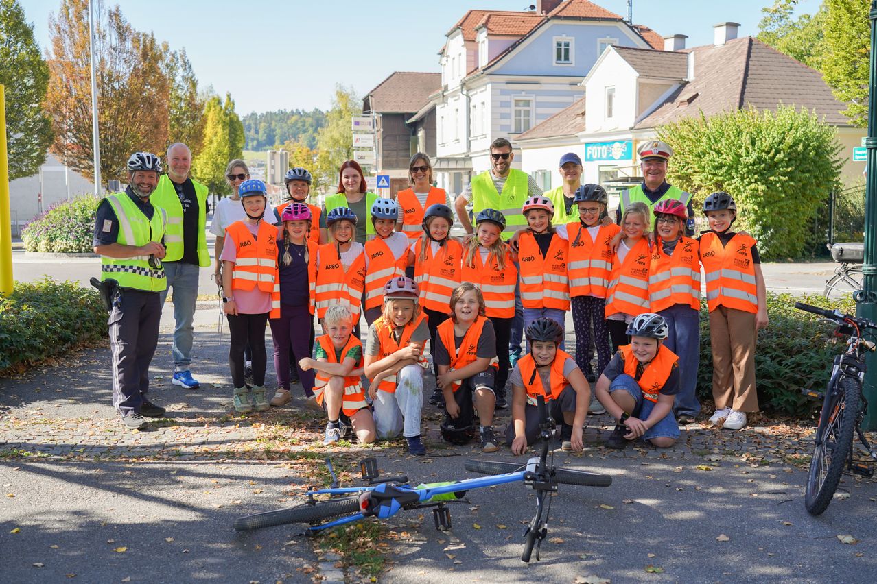Eine Gruppe von Kindern und Erwachsenen steht am Straßenrand, trägt orange Westen und Helme. Einige von ihnen halten Fahrräder.