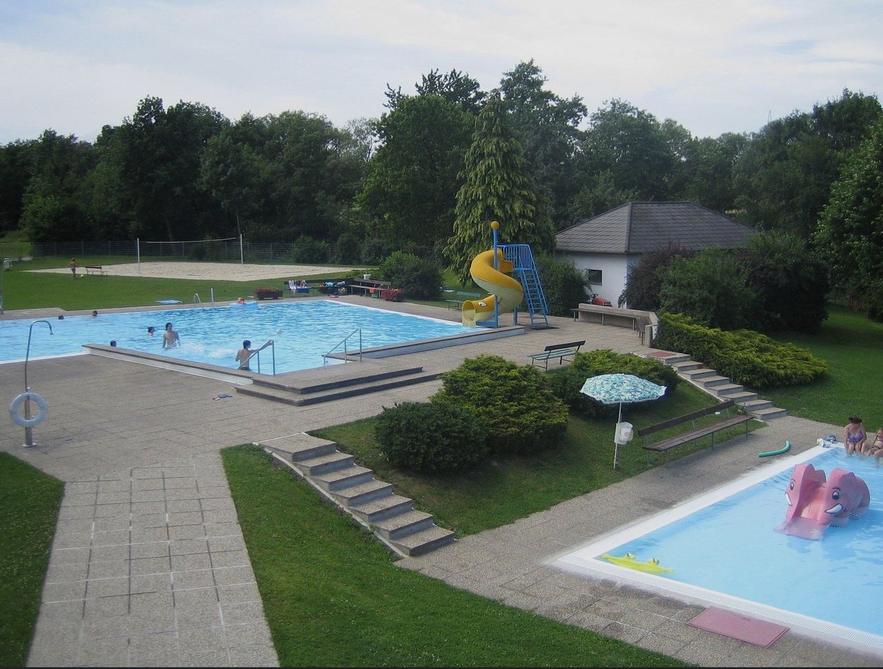 An aerial view of a pool area with people swimming and a yellow water slide. A bench and an umbrella are on the grass. A house and a volleyball net are in the distance.