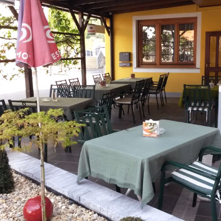 Ein Restaurant im Freien mit Tischen und Stühlen, grünen Tischdecken und einem roten Sonnenschirm. Das Gebäude hat gelbe Wände, Holzfenster und eine Glastür.