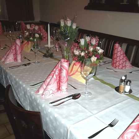 Ein langer Esstisch mit weißer Tischdecke und rosa Servietten, einer Kerze, Blumen und Besteck. Stühle sind um den Tisch herum angeordnet. Die Wand ist weiß.