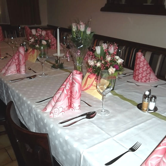 Ein langer Esstisch mit weißer Tischdecke und rosa Servietten, einer Kerze, Blumen und Besteck. Stühle sind um den Tisch herum angeordnet. Die Wand ist weiß.