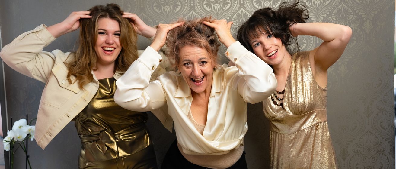 Drei Frauen in Metallic-Kleidung posieren für ein Foto. Die Frau in der Mitte hat ihre Haare in den Händen, während die anderen ihre Hände auf den Kopf halten.