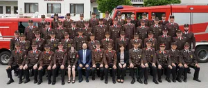 Eine Gruppe von Feuerwehrleuten in Uniform posiert für ein Foto vor einem Feuerwehrauto und einem Gebäude mit dem Schild Gemeindeamt.