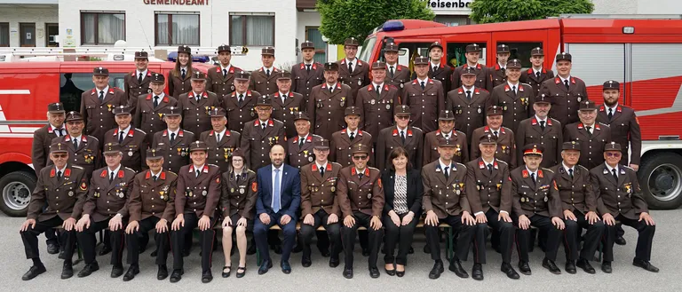 Eine Gruppe von Feuerwehrleuten in Uniform posiert für ein Foto vor einem Feuerwehrauto und einem Gebäude mit dem Schild Gemeindeamt.