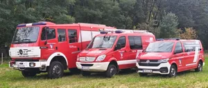 Drei rote Notfallfahrzeuge sind in einem Grasbereich mit Bäumen im Hintergrund geparkt.