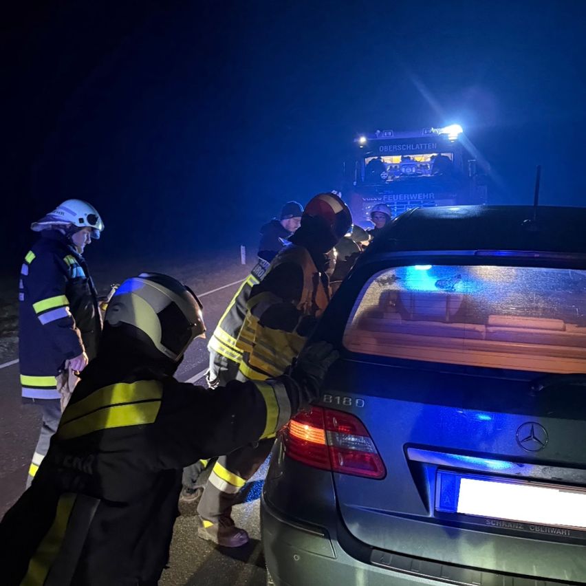 Einsatzkräfte in Schutzkleidung helfen bei einem Fahrzeug am Straßenrand in der Nacht, wobei ein Feuerwehrwagen dahinter geparkt ist.