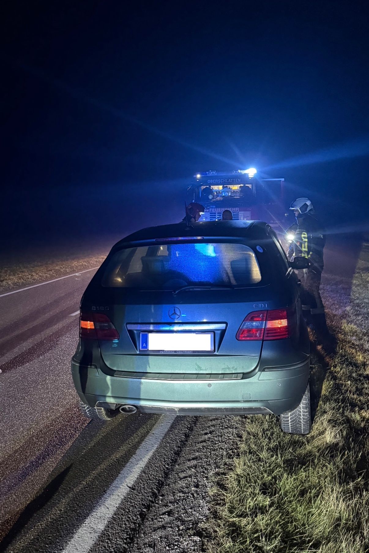 Ein blauer Mercedes-Wagen ist nachts am Straßenrand geparkt. Ein Feuerwehrmann steht mit einer Taschenlampe hinter dem Auto. Eine weitere Person befindet sich in der Nähe des Lastwagens, dessen Lichter eingeschaltet sind.