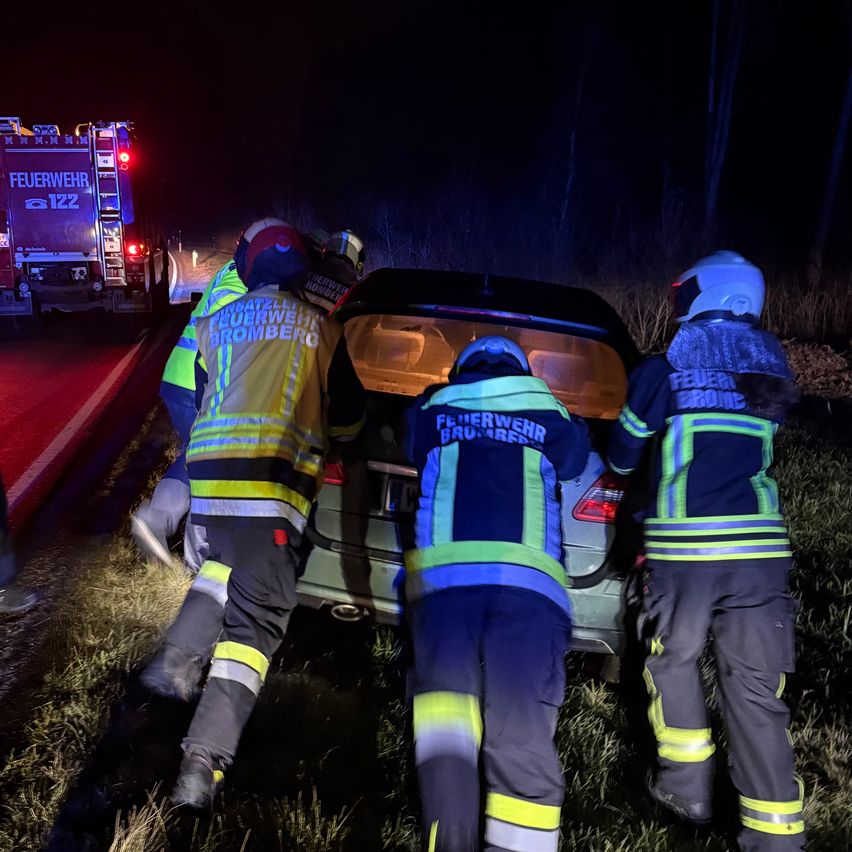 Feuerwehrleute in voller Ausrüstung stehen bei Nacht am Straßenrand und heben die Rückseite eines Autos an. Sie tragen reflektierende Westen mit 'Feuerwehr' und 'Bromberg' darauf. Ein Feuerwehrwagen ist hinter ihnen geparkt.