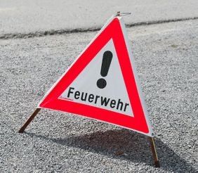 Ein rotes und weißes Dreiecksschild mit einem Ausrufezeichen und dem Wort 'Feuerwehr' darauf, das wahrscheinlich auf die Nähe einer Feuerwehr hinweist.