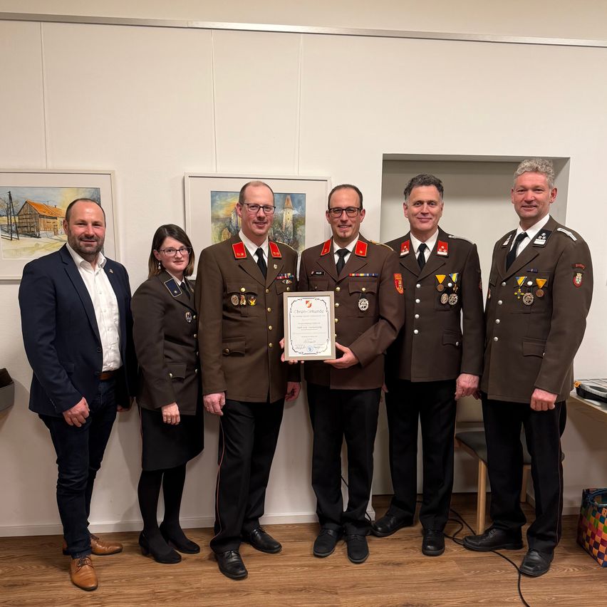 Eine Gruppe uniformierter Personen steht in einem Raum und überreicht einem zentralen Akteur eine Urkunde. Zwei Personen tragen Anzüge, und drei tragen Militäruniformen. Dahinter hält eine weiße Wand gerahmte Gemälde.