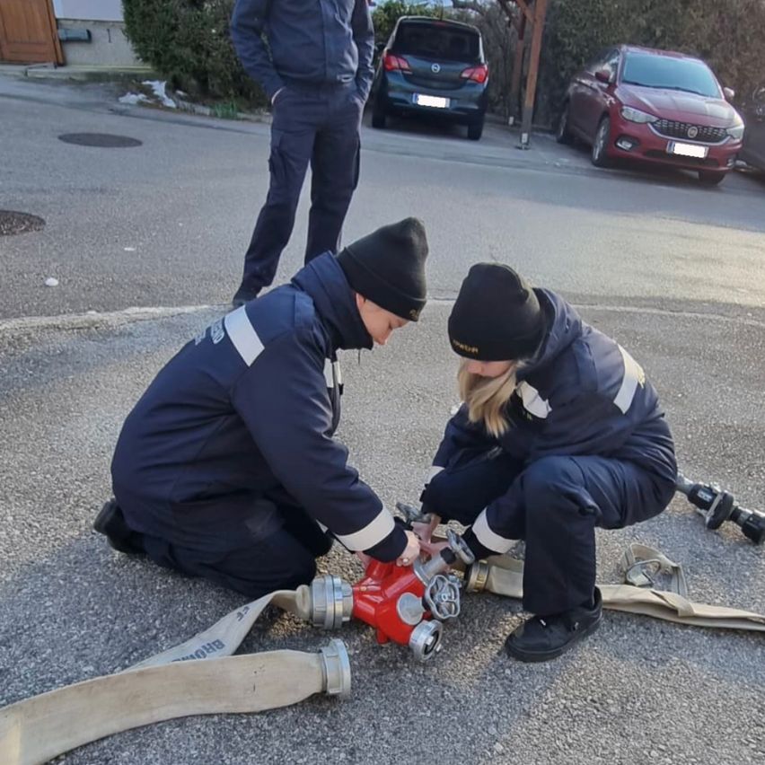 Zwei Feuerwehrleute sind auf der Straße, einer von ihnen kniet und arbeitet an einem Feuerhydranten, während der andere in der Nähe steht, mit zwei geparkten Autos dahinter.