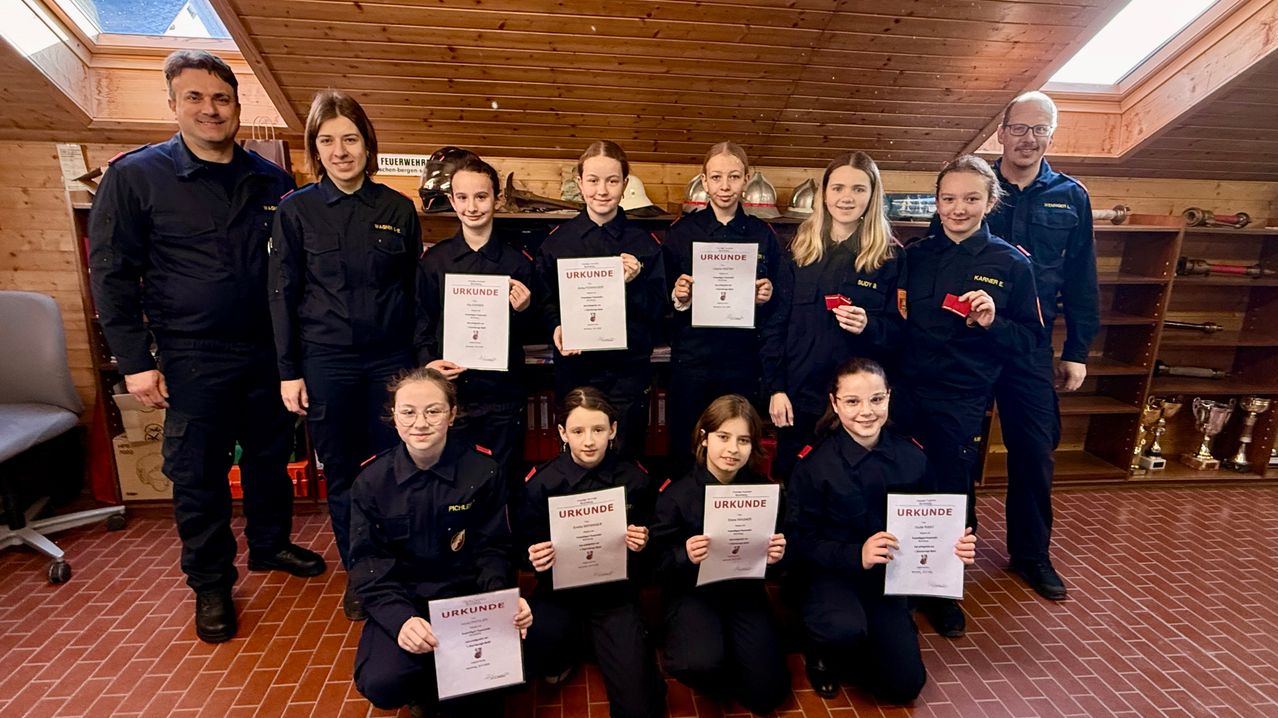Eine Gruppe junger Frauen in identischen Uniformen posiert für ein Foto und hält Urkunden mit der Aufschrift 'Urkunde'. Sie stehen in einem Raum mit Holzwänden und einem Ziegelboden.