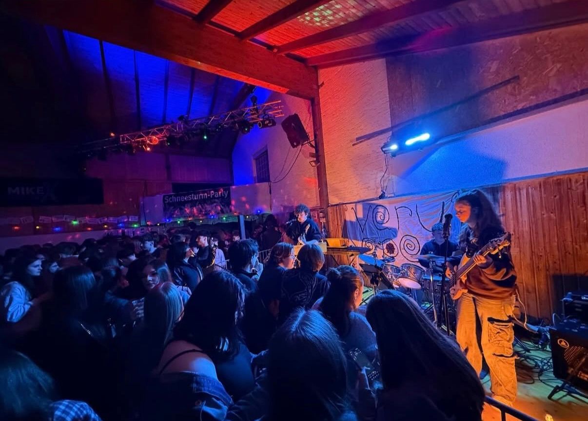 Eine Band spielt auf der Bühne in einem farbenfroh beleuchteten Veranstaltungsort. Zwei Frauen spielen Gitarre, während andere Schlagzeug und Keyboard spielen. Das Publikum, beleuchtet von Bühnenlichtern, sitzt vor der Band.