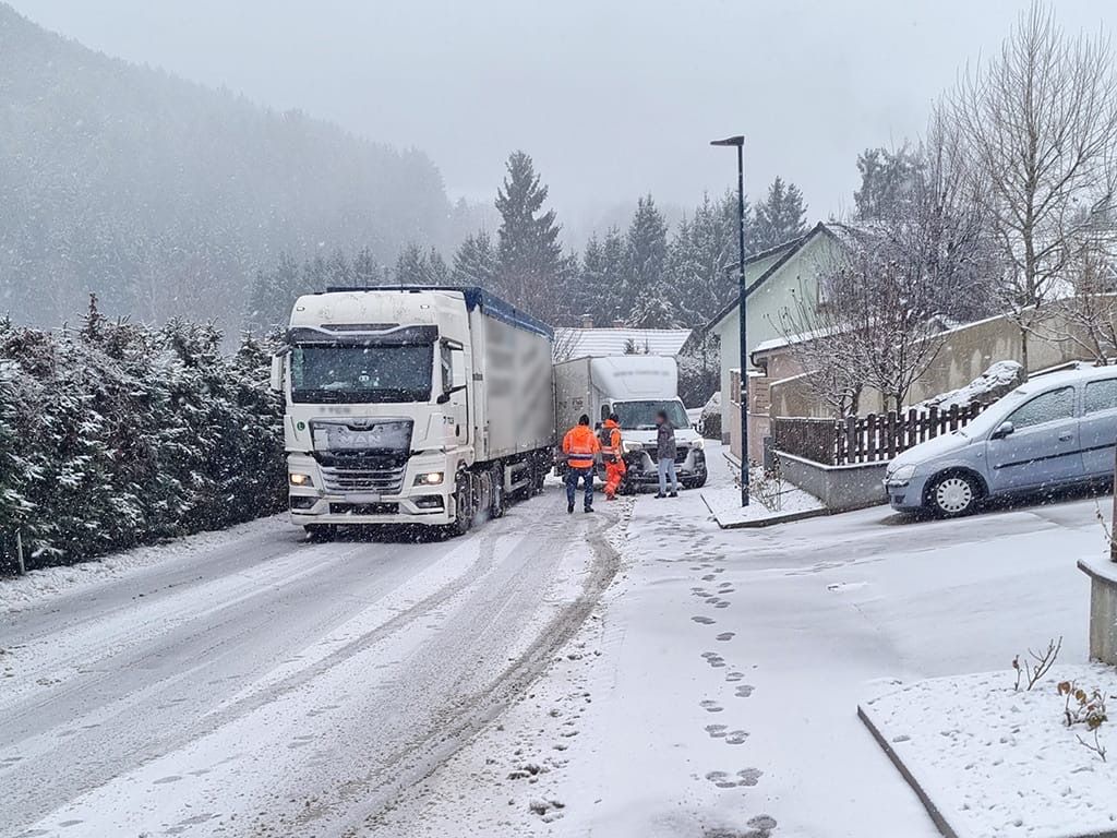 Eine verschneite Straße mit einem Lastwagen, der darauf fährt. An der Seite stehen ein Mann in einer orangefarbenen Weste und ein weiterer in Grau. In der Nähe befinden sich ein Zaun und ein geparktes Auto.