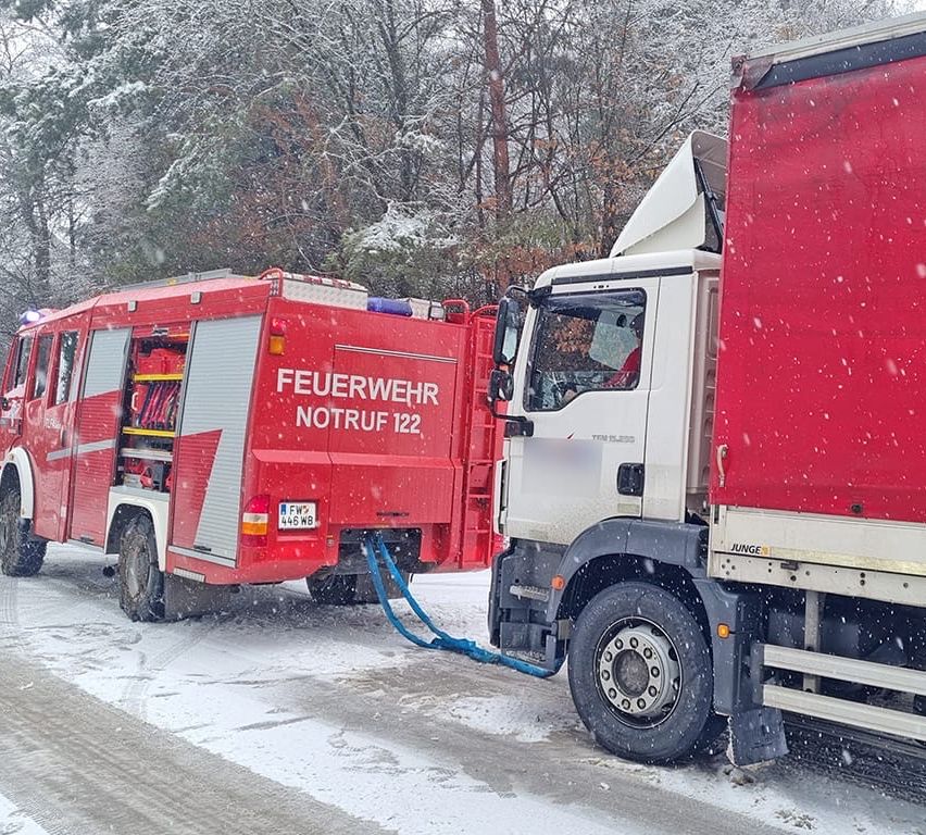 Ein roter Feuerwehrwagen mit der Aufschrift 'FEUERWEHR NOTRUF 122' steht auf verschneiter Straße, neben einem Lastwagen. Eine Feuerwehrschlauch ist angeschlossen.