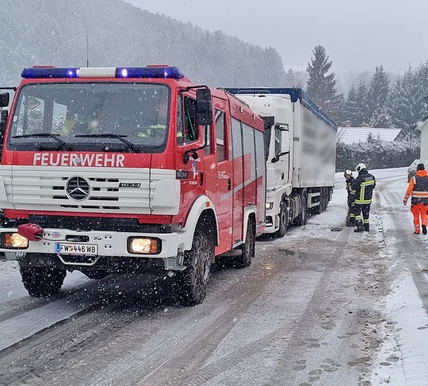 Ein roter Feuerwehrwagen mit dem Wort 'Feuerwehr' auf der Front fährt auf einer verschneiten Straße, mit Feuerwehrleuten in der Nähe. Dahinter steht ein weißer Lastwagen mit verschwommenen Details. Bäume und ein Haus sind im Hintergrund zu sehen.