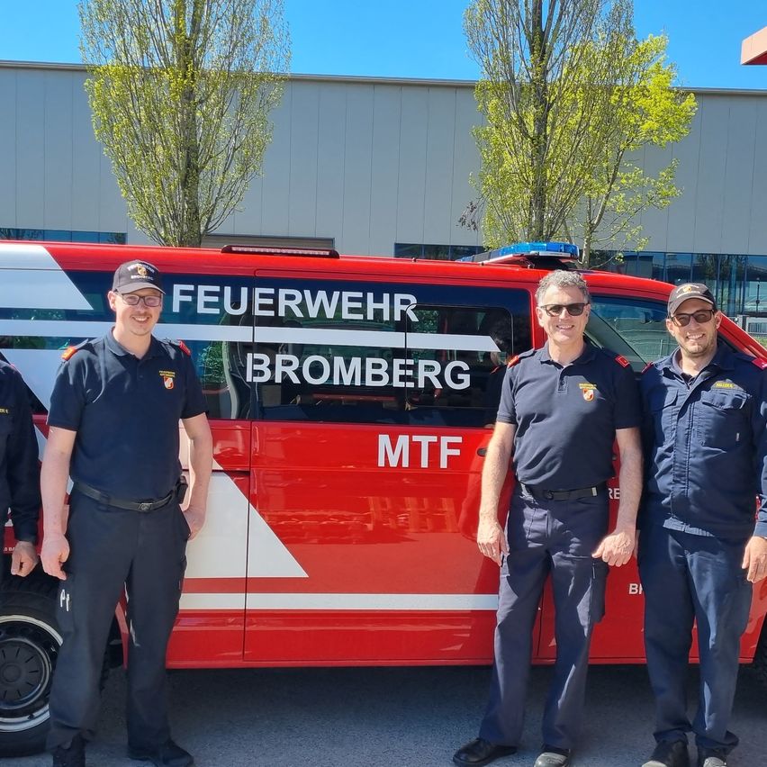 Three firefighters stand in front of a red vehicle with the word Bromberg on it. They are wearing uniforms and hats.