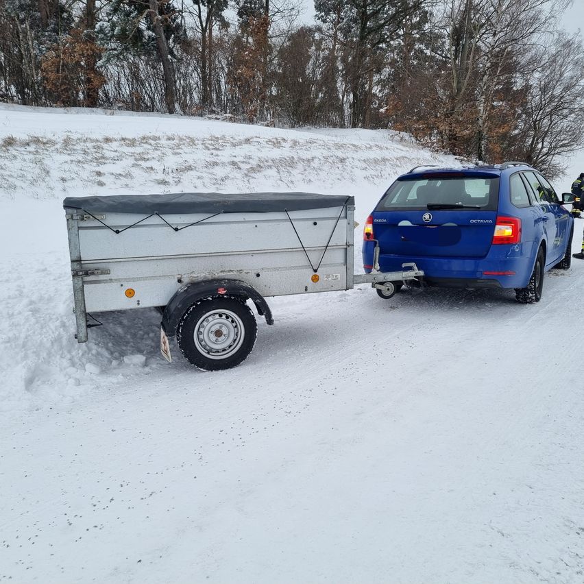 A blue car is towing a white trailer in a snowy landscape. Behind them, a person in a yellow vest stands on the road.
