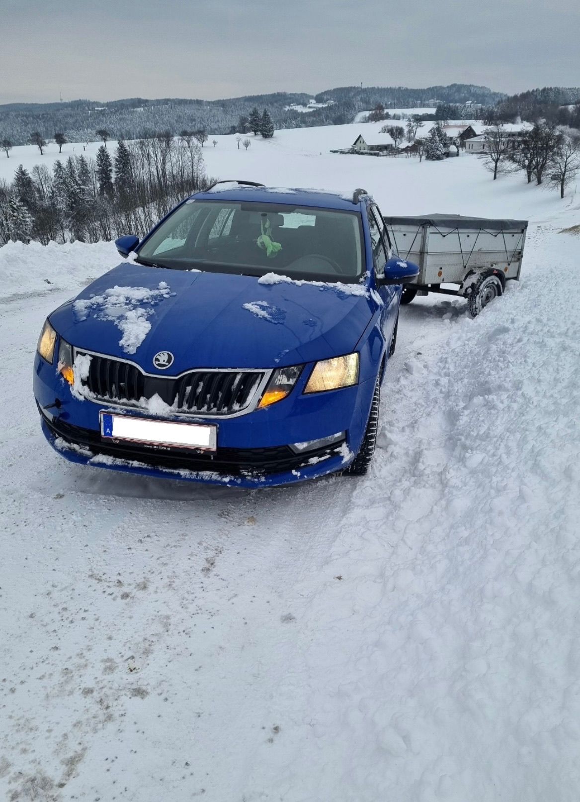 A blue Skoda car towing a trailer is stuck in snow on a road, with snow-covered trees and houses in the background.
