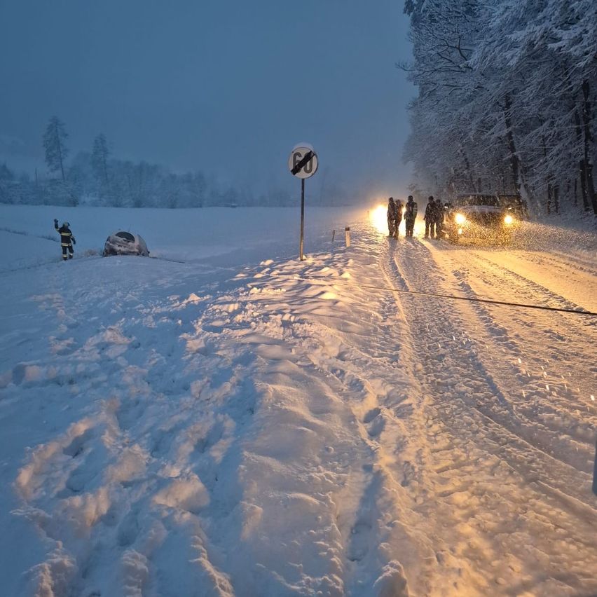People in winter gear assist a vehicle stuck in snow. Nearby, a sign indicates a no-entry zone.