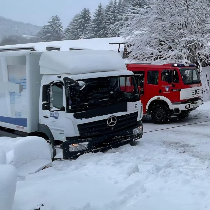 Two trucks, one white and one red, are parked on a snowy road. The white truck has a broken windshield and is partially buried in snow.