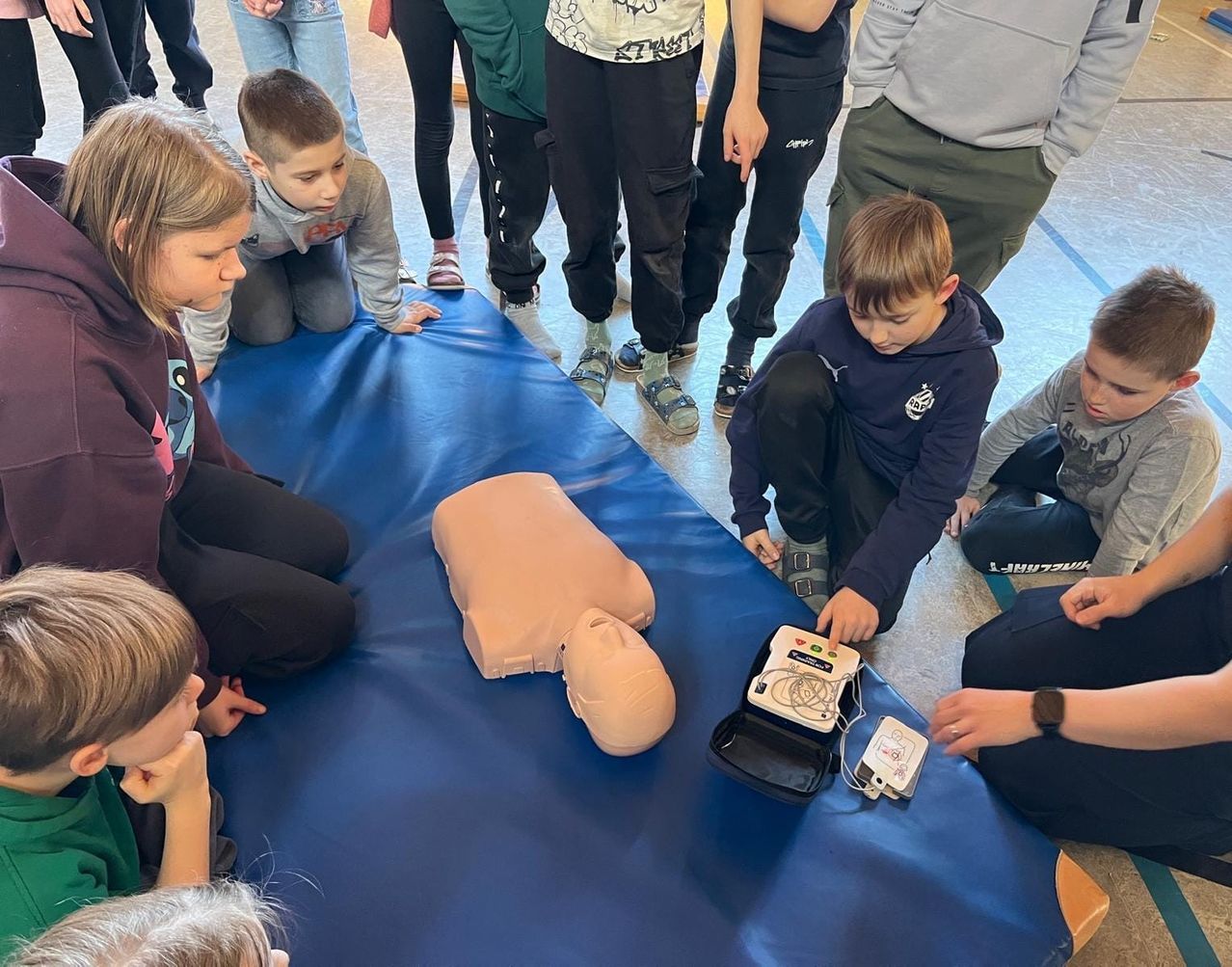 Kinder versammeln sich um eine CPR-Puppe auf einer blauen Matte, wobei ein Kind einen Defibrillator benutzt. Ein Lehrer unterrichtet sie in Erster Hilfe.