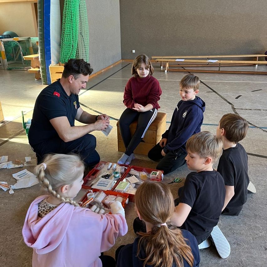 Ein Mann in Uniform unterrichtet Kinder über Erste Hilfe, während sie in einem Kreis um einen Erste-Hilfe-Kasten sitzen.