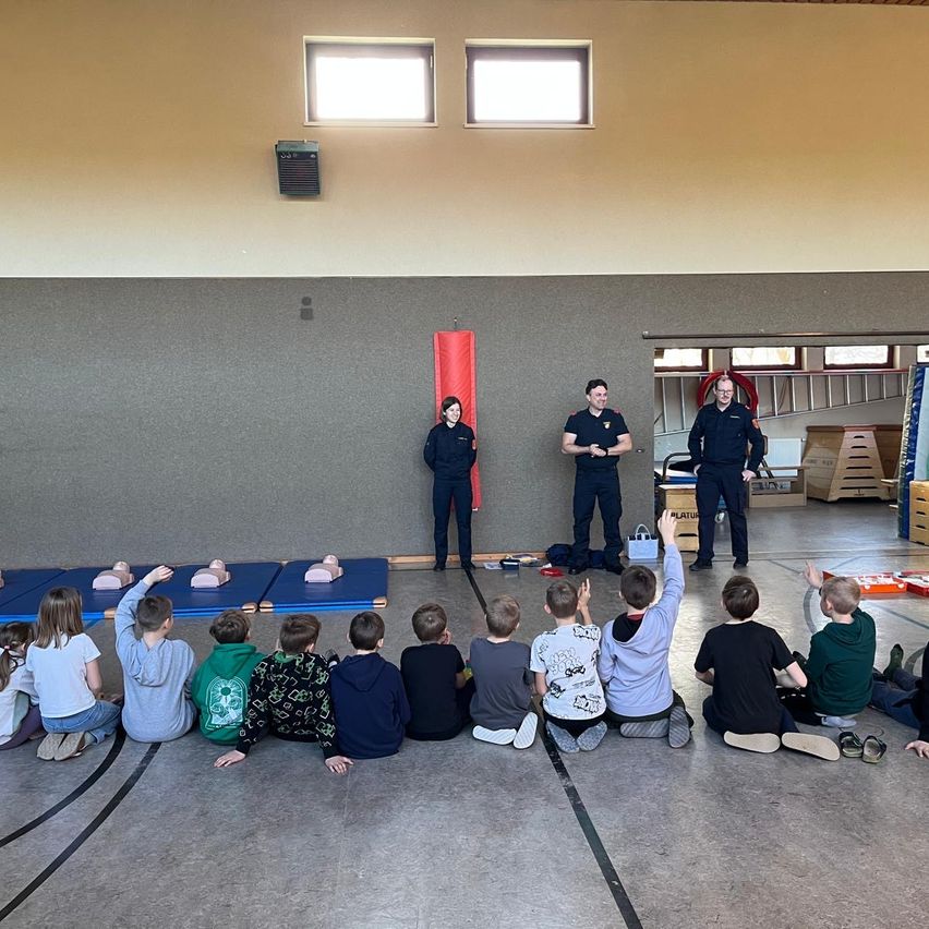Eine Gruppe von Kindern sitzt auf dem Boden in einem Raum und hört einem Mann in Uniform zu. Drei Männer stehen vor ihnen, mit CPR-Trainingspuppen auf blauen Matten hinter ihnen.