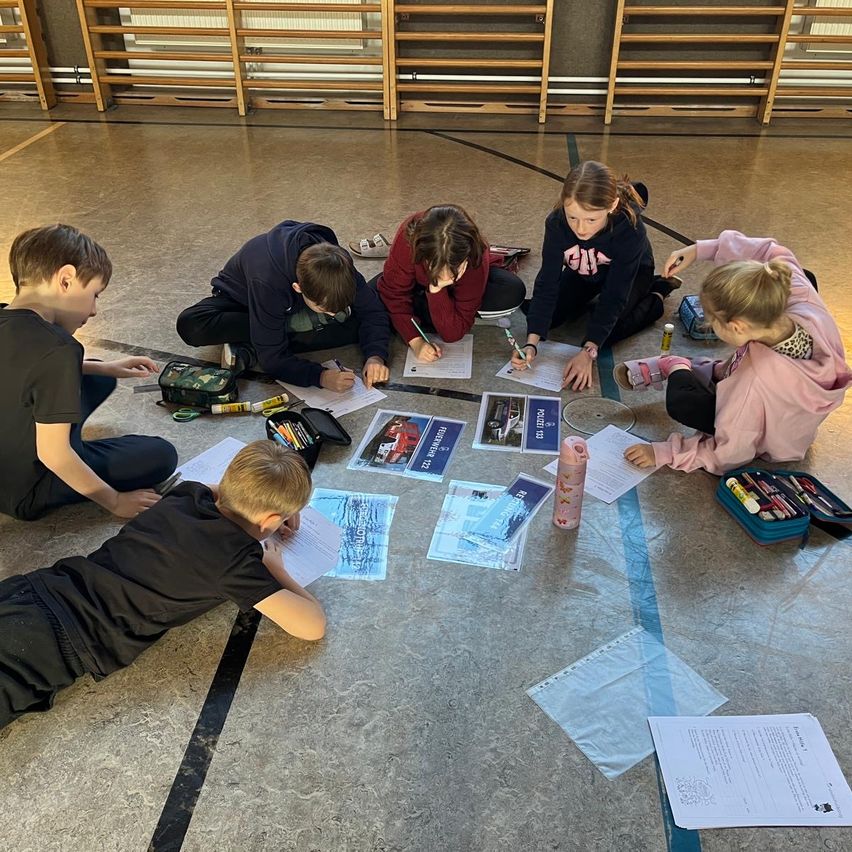 Eine Gruppe von Kindern sitzt in einem Kreis auf dem Boden, möglicherweise bei einer Schulaktivität. Sie haben Papiere, Stifte und andere Gegenstände auf dem Boden. Sie tragen alle Freizeitkleidung. Der Boden hat eine schwarze Linie. Hinter ihnen steht eine Holzstruktur.