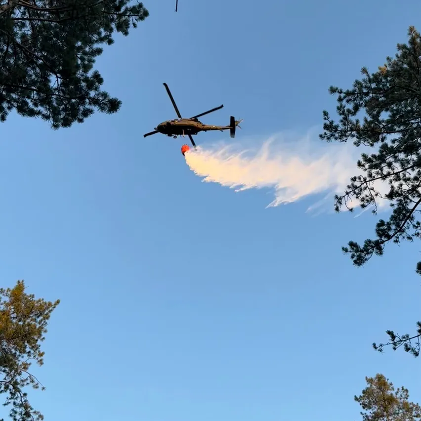 Ein Hubschrauber fliegt über einen Wald und gibt Wasser ab, um ein Feuer zu löschen.