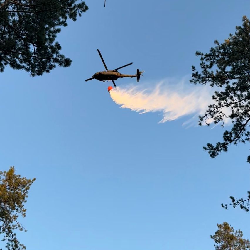 A helicopter is flying over a forest, releasing water to extinguish a fire.