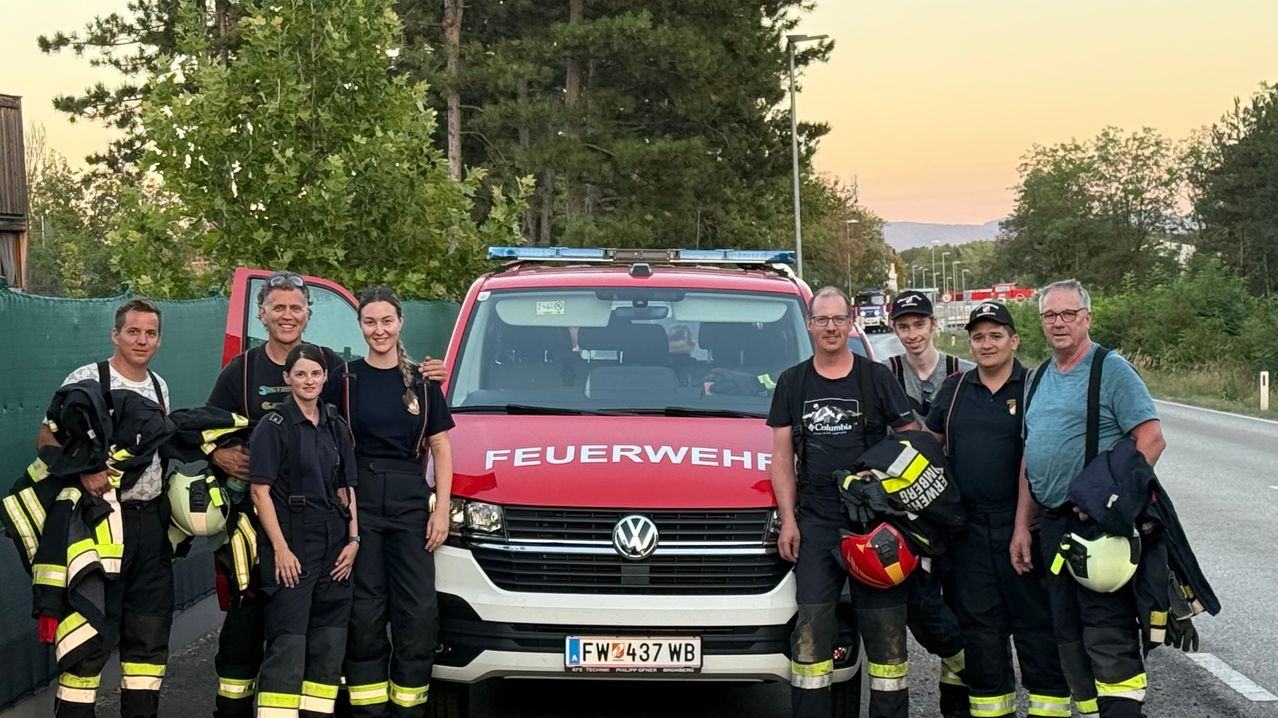A group of firefighters in uniform stand next to a red fire truck with the word 'Feuerwehr' on it. They are posing for a photo.