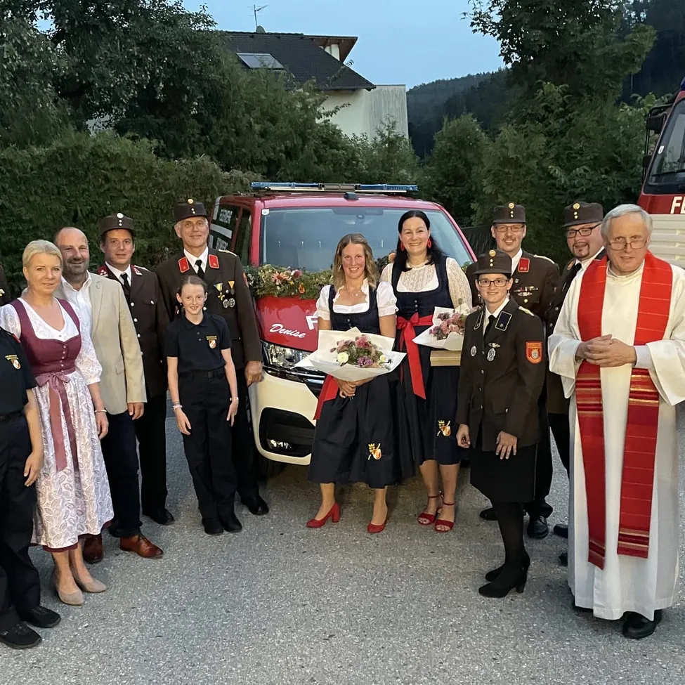 Eine Gruppe von Menschen in traditioneller Kleidung und Uniformen steht vor einem roten Fahrzeug, mit einem Priester rechts und einer Frau mit Blumen in der Mitte.