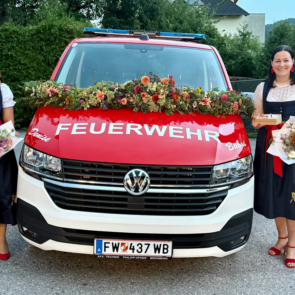 Zwei Frauen in traditioneller Kleidung stehen neben einem roten Feuerwehrauto mit Blumen auf der Front, halten Blumensträuße. Das Wort 'Feuerwehr' steht auf dem Auto.