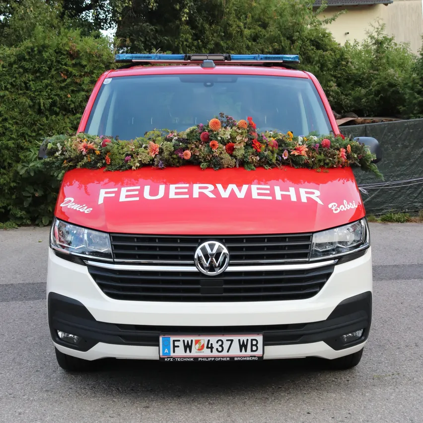 Ein roter und weißer Volkswagen-Transporter mit der Aufschrift 'FEUERWEHR', geschmückt mit Blumen auf der Vorderseite. Das Kennzeichen lautet 'FW 437 WB'.