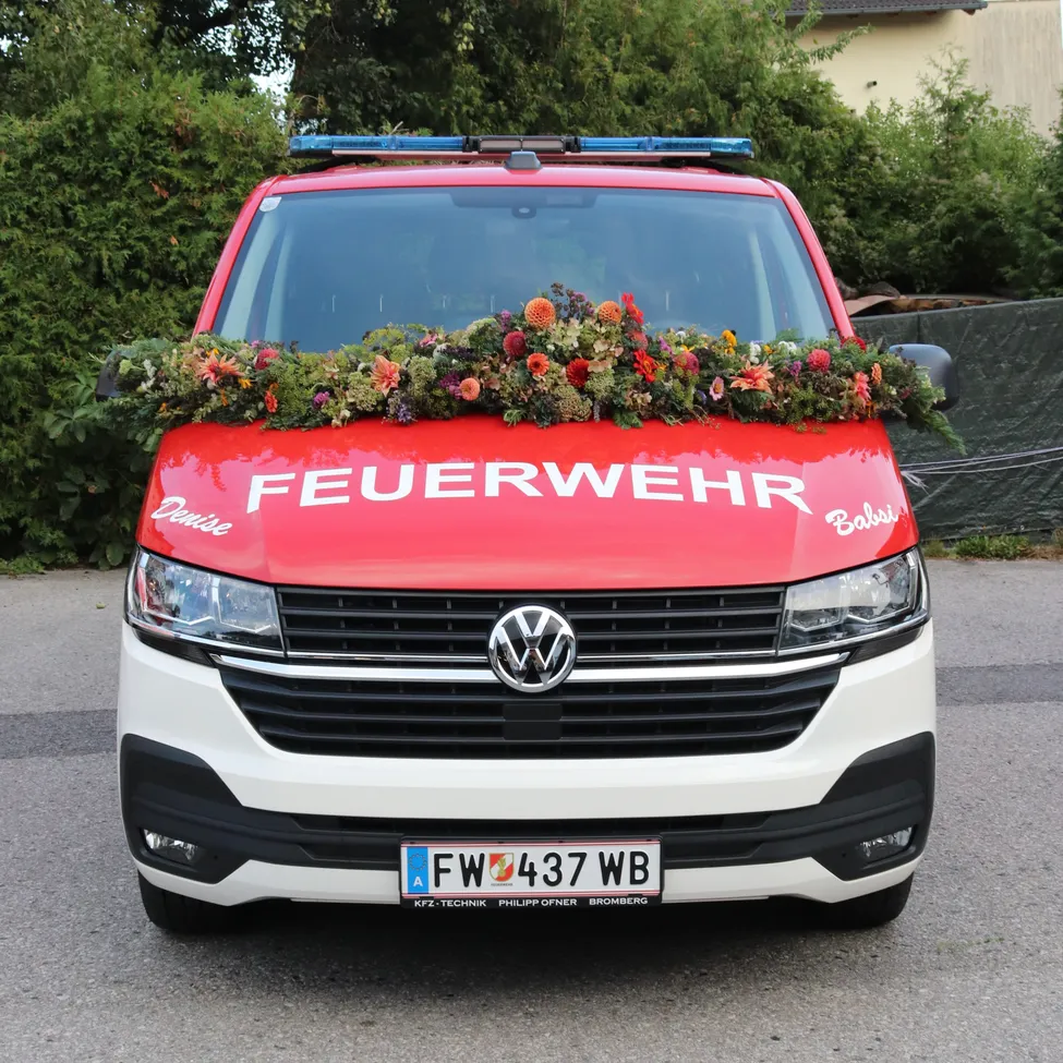 Ein roter und weißer Volkswagen-Transporter mit der Aufschrift 'FEUERWEHR', geschmückt mit Blumen auf der Vorderseite. Das Kennzeichen lautet 'FW 437 WB'.