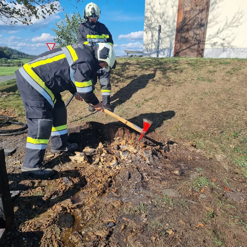 Zwei Feuerwehrleute in voller Montur arbeiten an einem schlammigen Ort, einer benutzt einen roten Axt zum Graben. Ein Gebäude mit einer Holztür ist im Hintergrund.