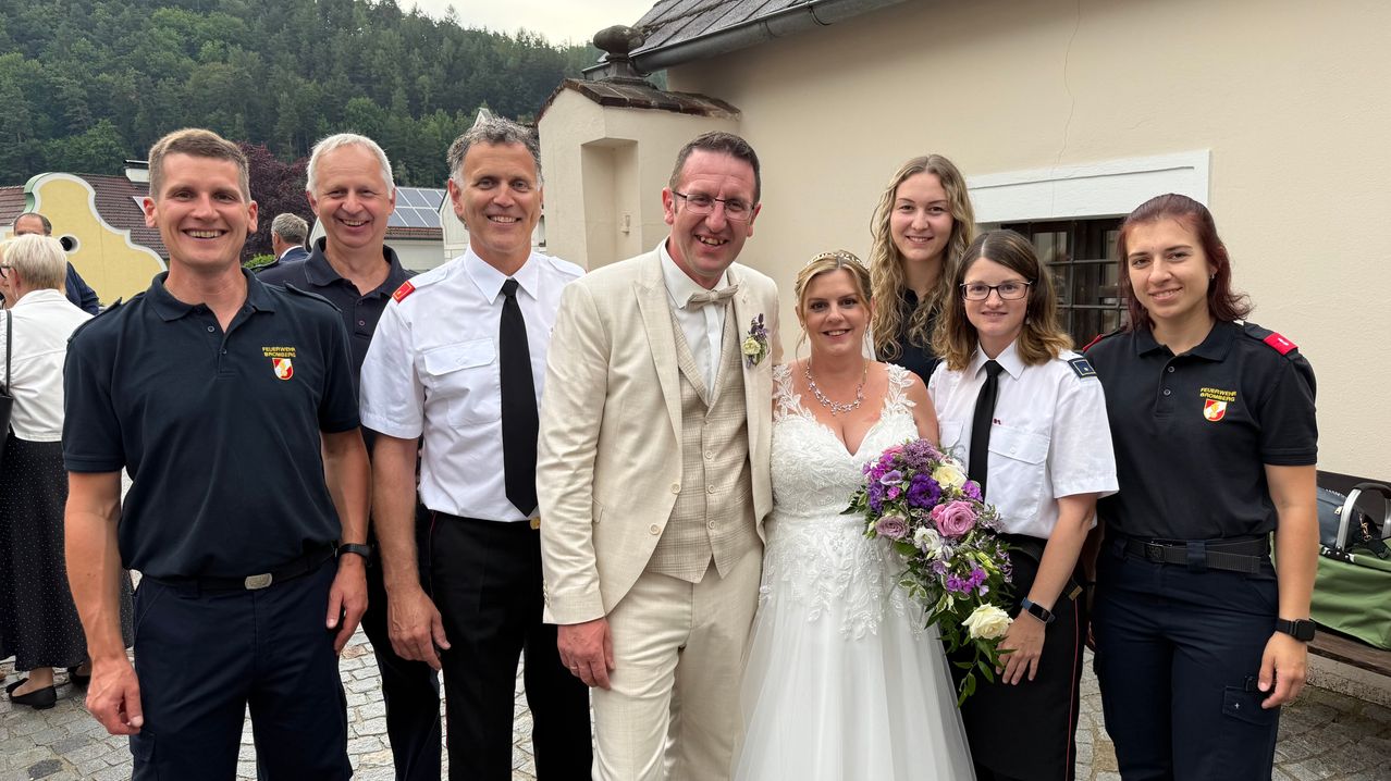 Eine Hochzeitsfeier vor einem Haus mit drei Frauen und drei Männern, die für ein Foto lächeln. Der Bräutigam trägt einen weißen Anzug mit Fliege. Die Braut im Hochzeitskleid hält einen Blumenstrauß.