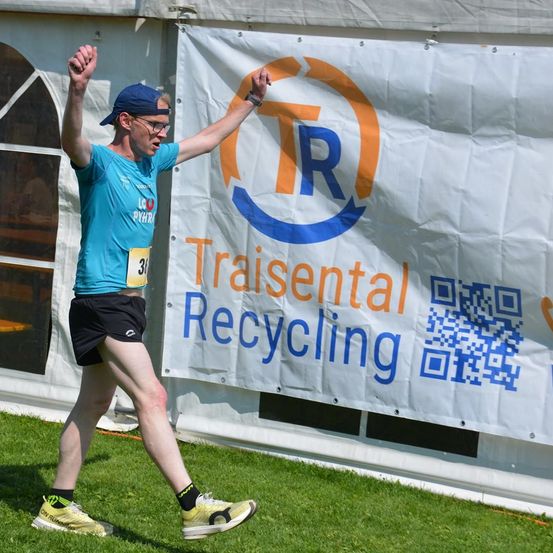 Ein Mann in einem blauen Shirt und schwarzen Shorts läuft einen Marathon. Hinter ihm befindet sich ein weißer Banner mit den Worten 'Traisental Recycling' und einem Logo.