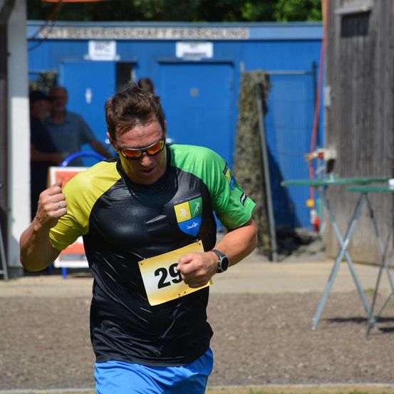 Ein Mann läuft in einem Rennen und trägt eine Nummer. Er trägt eine grüne, gelbe und schwarze Kleidung. Hinter ihm steht ein blaues Gebäude und einige Zuschauer.